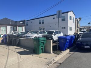 Recycling and trash bins lined up outside residential buildings on a sunny day.